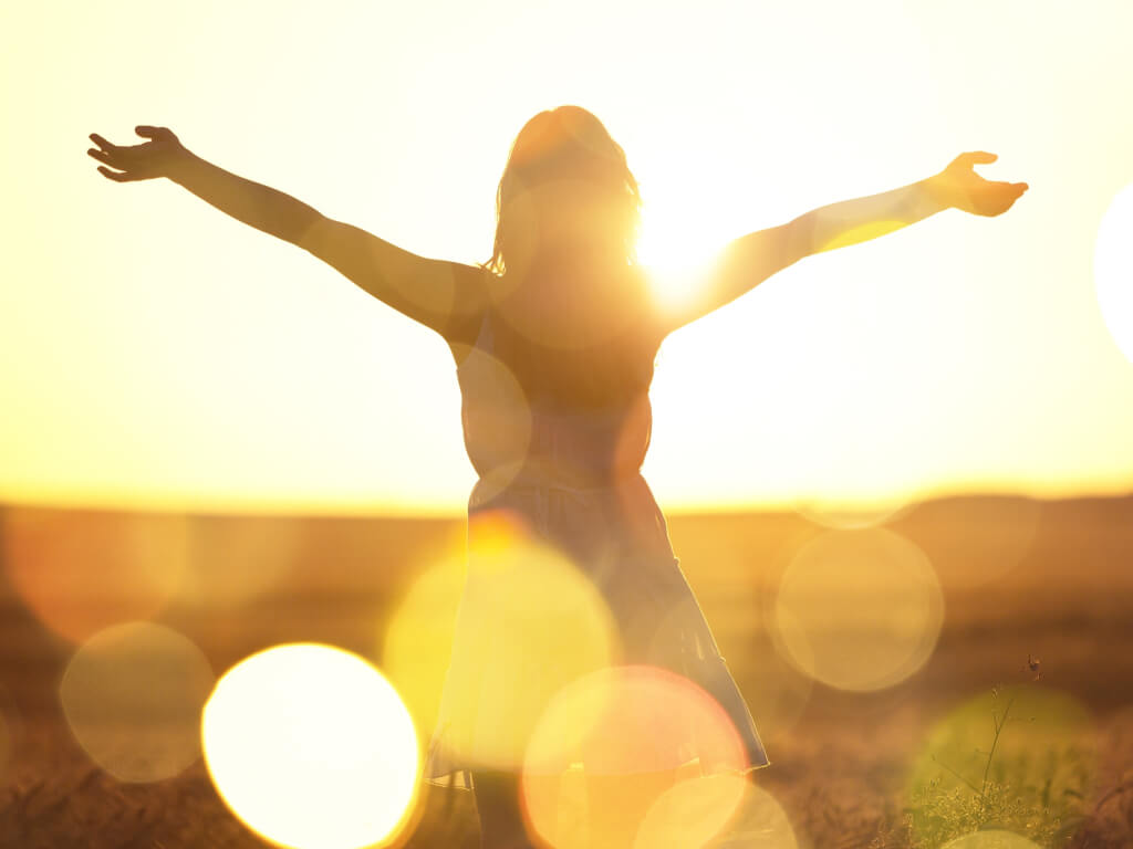 Sunlit silhouette of a woman with outstretched hands standing in field